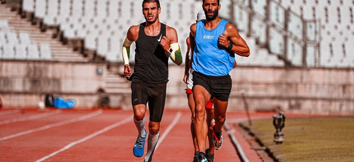 Two male runners competing on a stadium track during a sunny day.