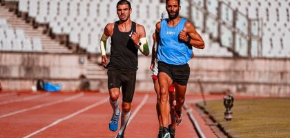 Two male runners competing on a stadium track during a sunny day.