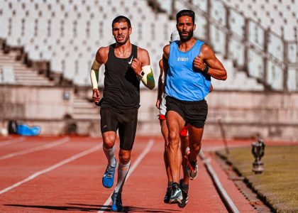 Two male runners competing on a stadium track during a sunny day.